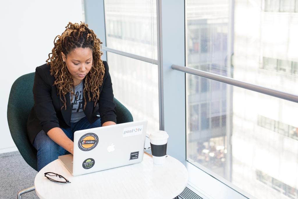 A Black woman with long, curly hair in locs, wearing jeans, a tee shirt, and a black blazer, works at a laptop next to a bright, sunny window with office buildings outside.